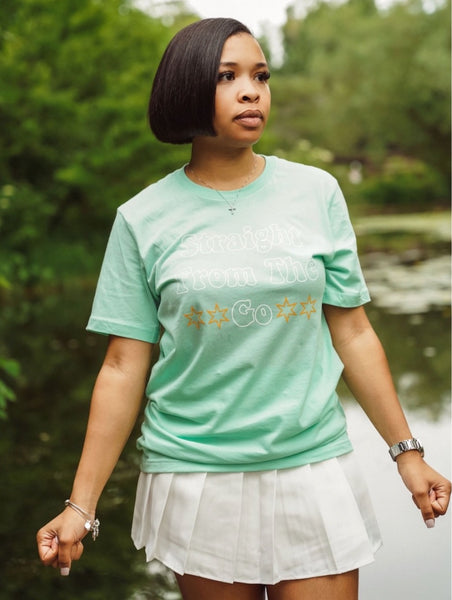 Black Woman wearing a mint green SFTG tee shirt in a white tennis skirt standing in front of a lake outlined by trees .